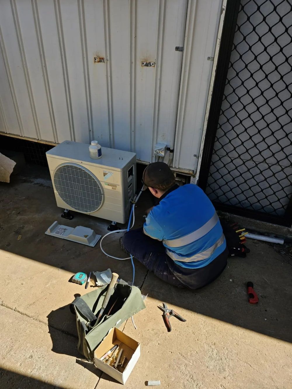 A Person in Blue Works on an Ac Unit Near a Corrugated Wall — BNS Refrigeration Services in Healy, QLD