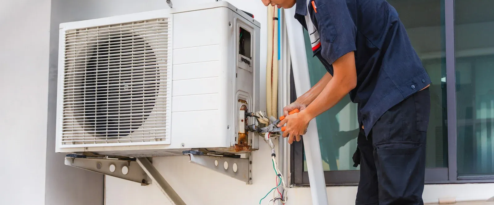 Technician servicing an outdoor air conditioner unit with tools near a building exterior