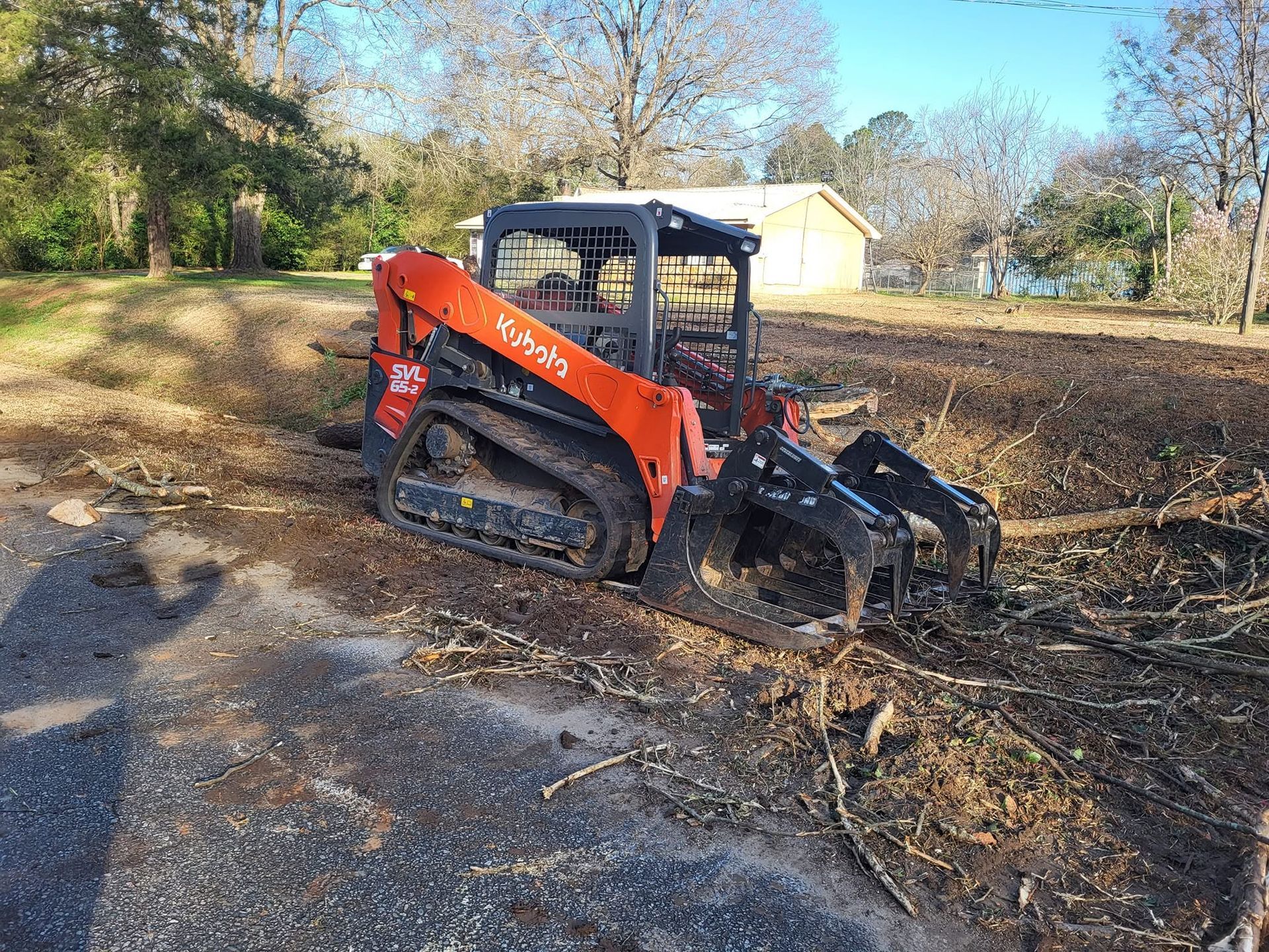 Orange skid steer clearing muddy debris on a dirt path near a house and trees