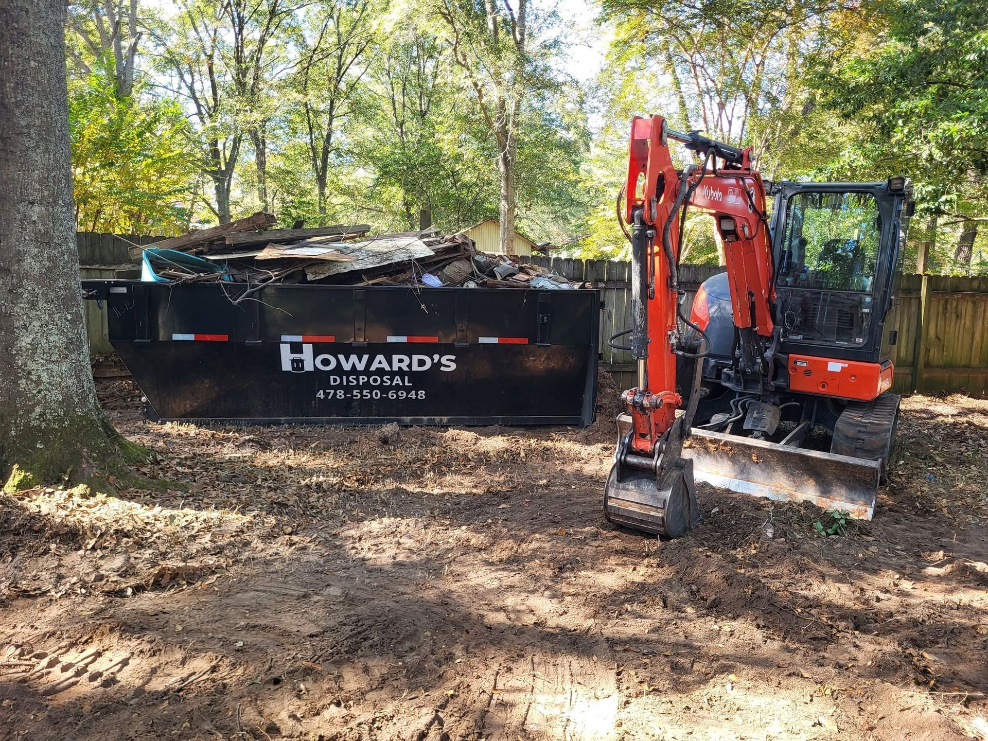 Red excavator beside a Howard’s dumpster in a wooded worksite covered with leaves.