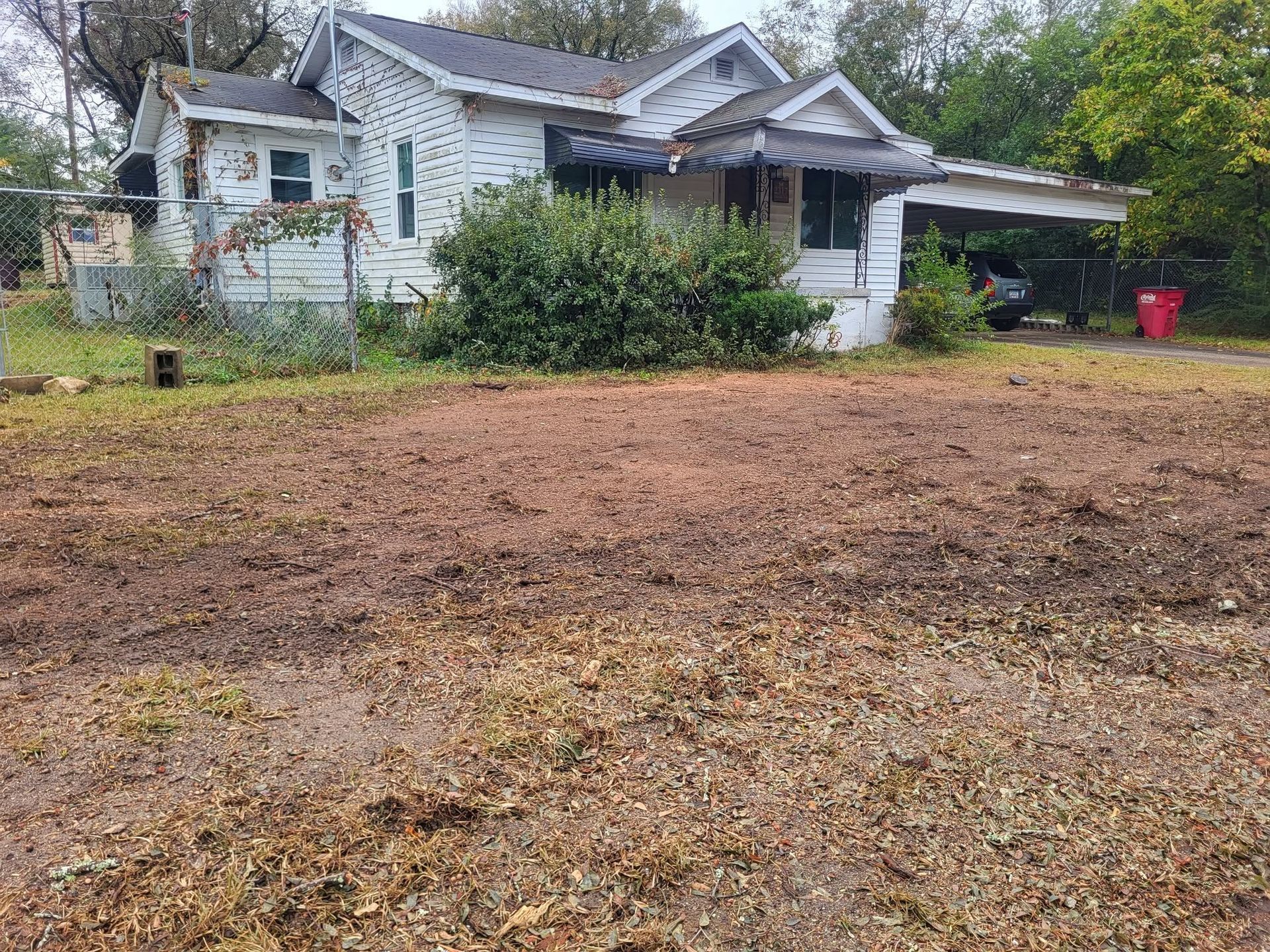 Overgrown yard in front of an old white house with a carport and red trash bin