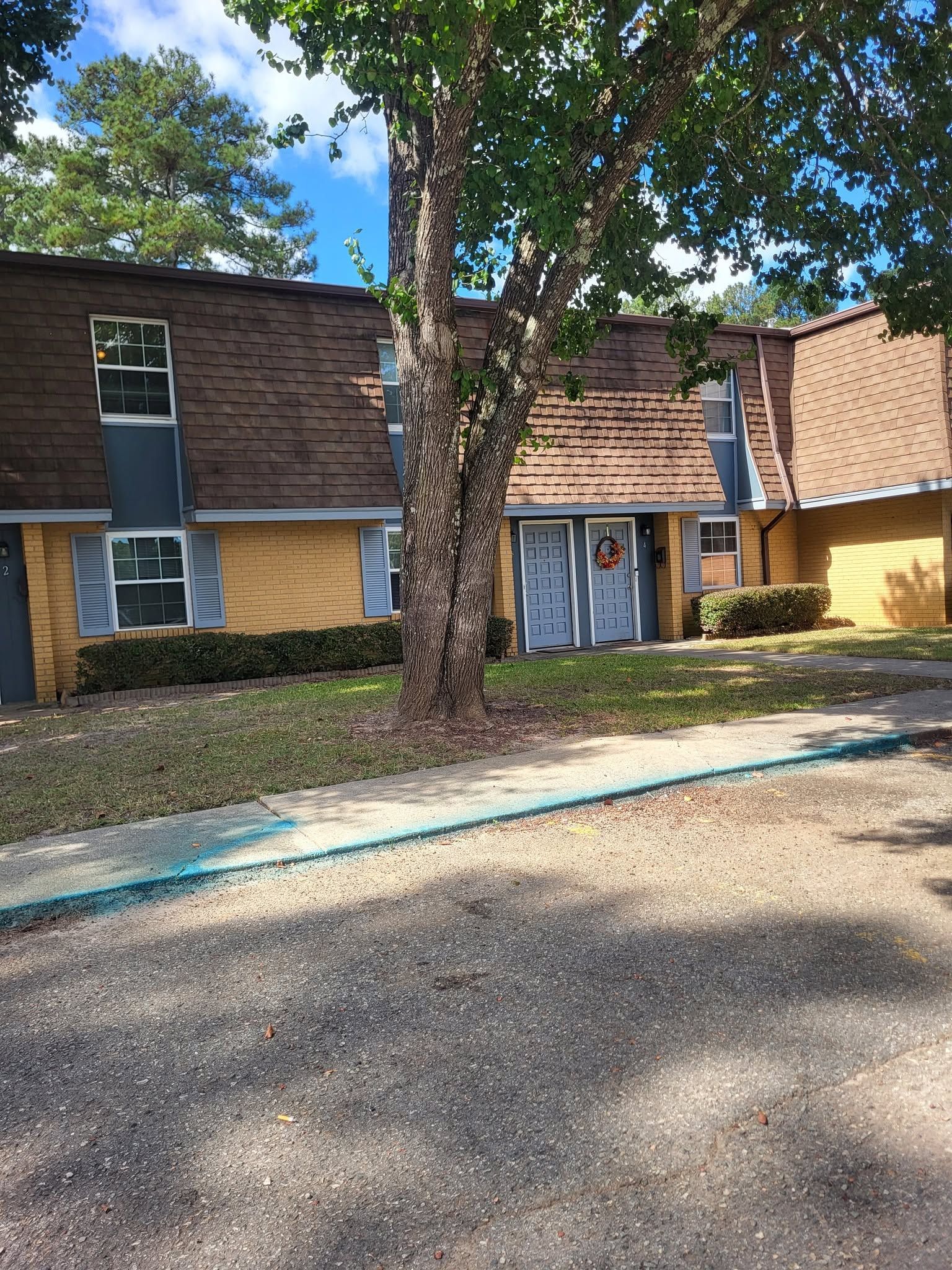 Apartment building with brown brick, blue doors, and a large tree in front by a gravel driveway.