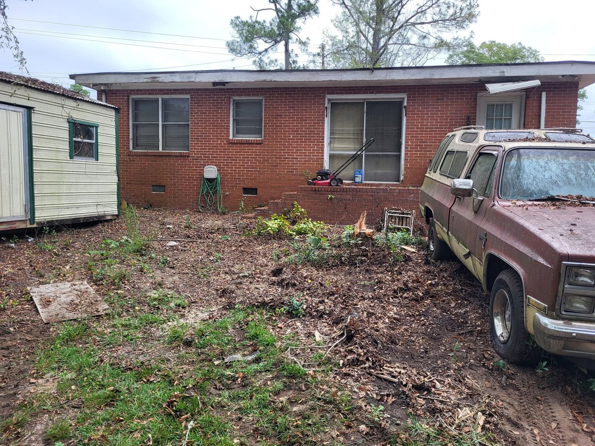 Overgrown yard beside a brick house, with an old brown SUV parked on the right.
