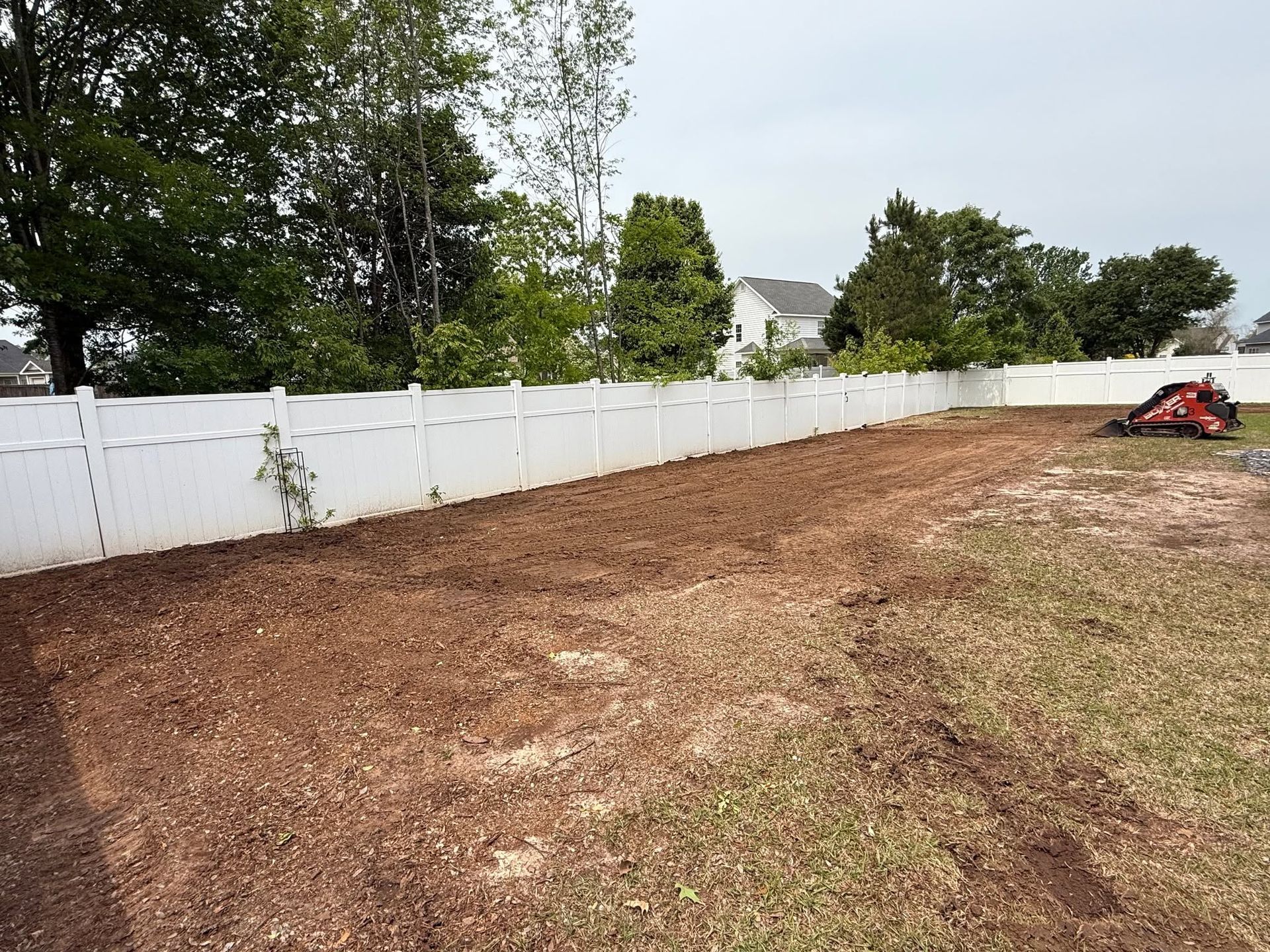 Bare backyard with fresh dirt, white vinyl fence, trees, and a red lawn tractor at the far right.