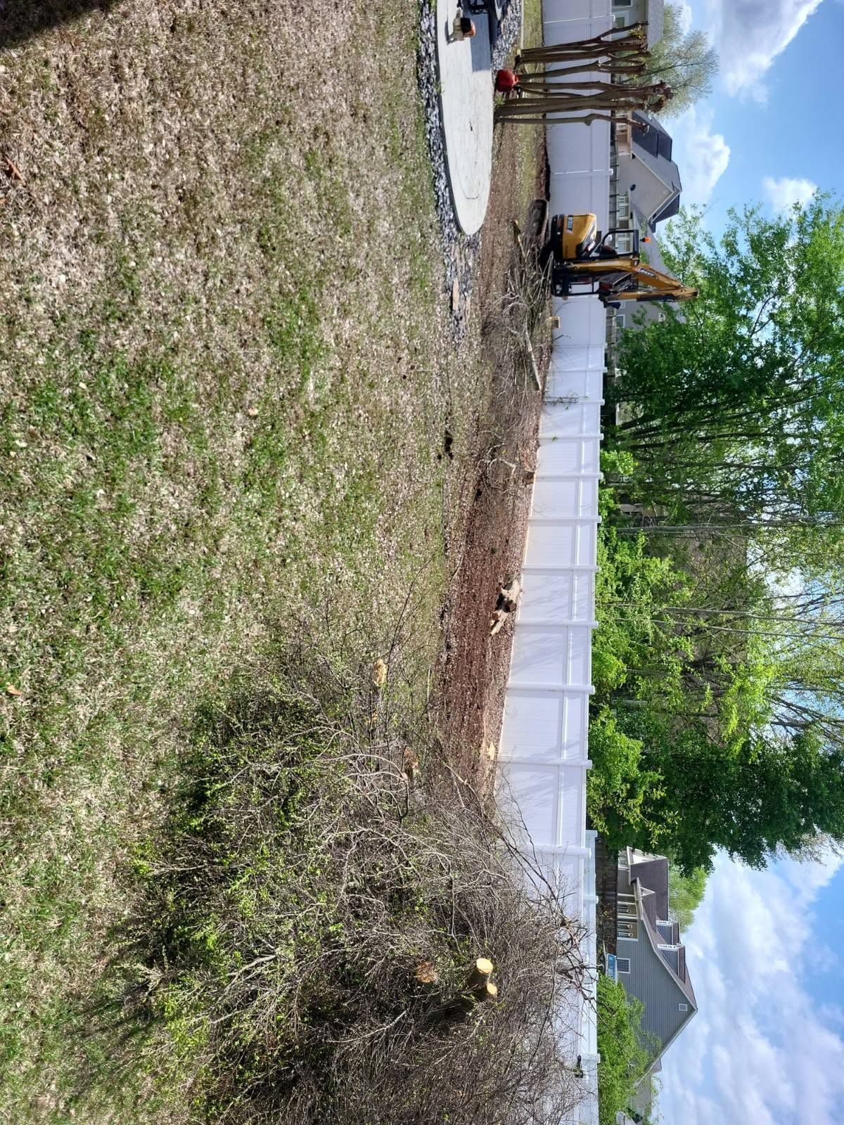 Construction site with excavator beside a white fence and dirt lot under a cloudy sky