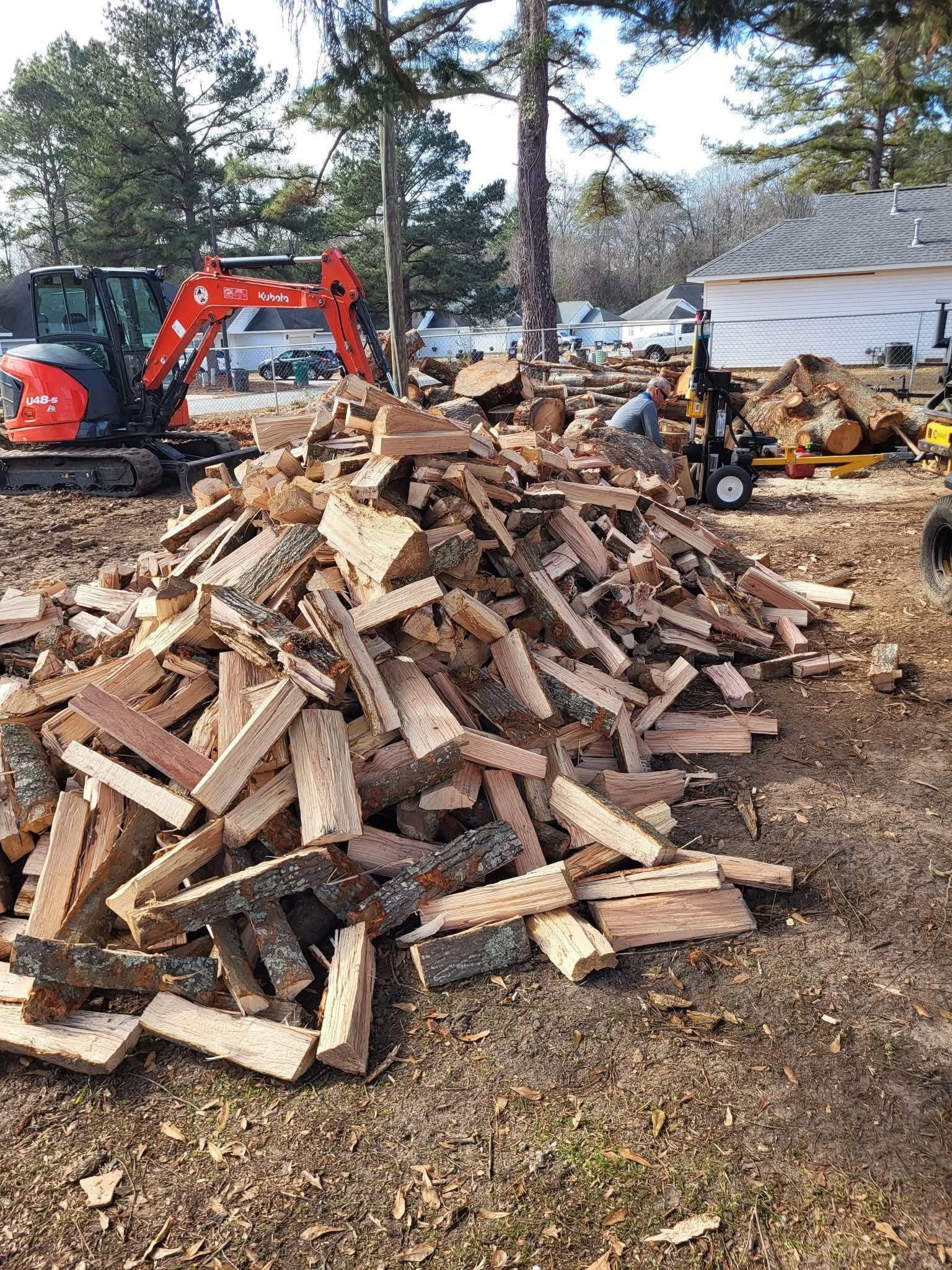 Orange excavator beside a large pile of wooden debris at a demolition site, with buildings in the background