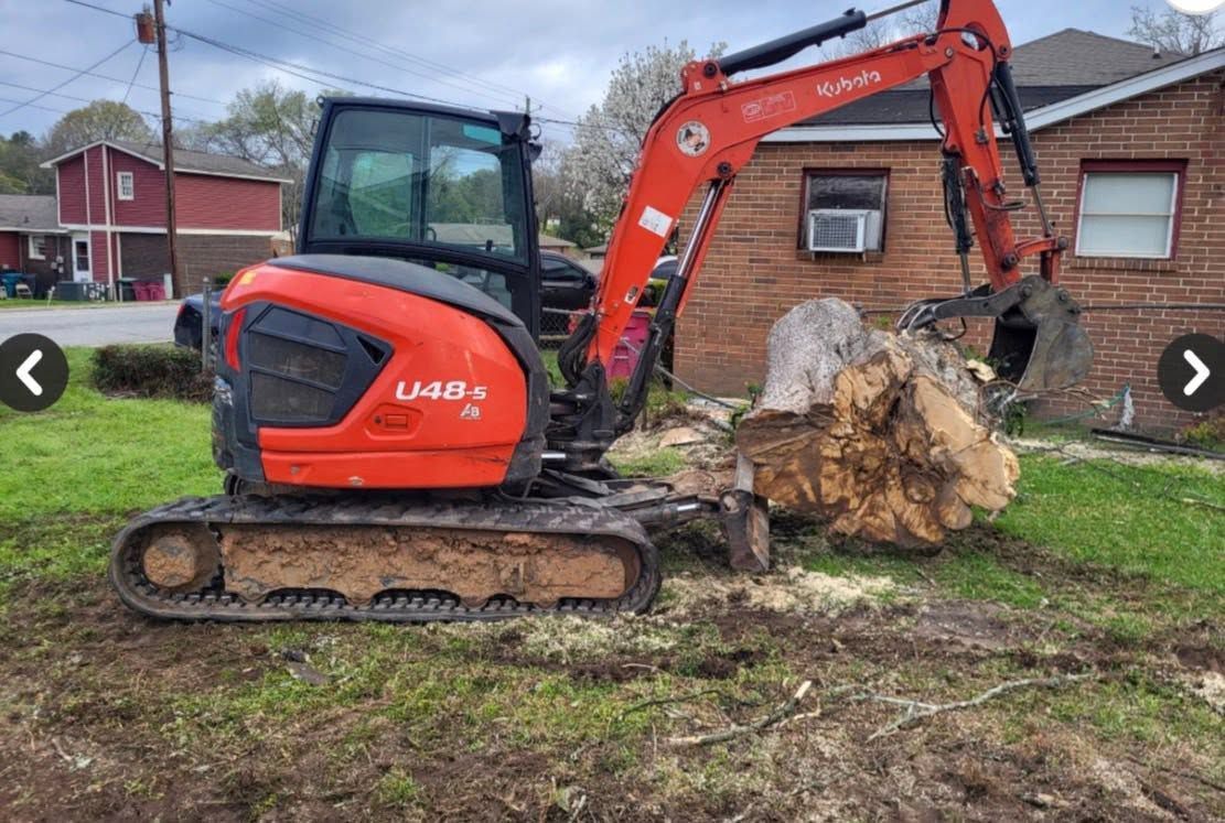 Orange excavator lifting a large tree stump beside a brick house on a grassy yard