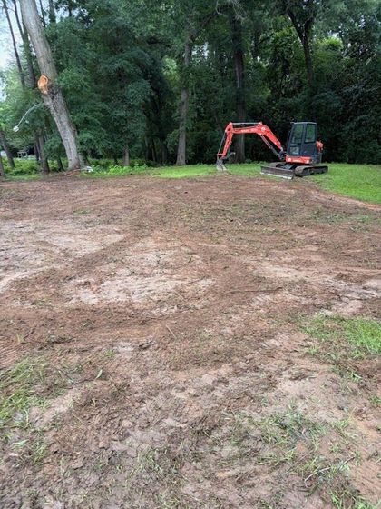 Cleared dirt lot beside trees with a red mini excavator in the background