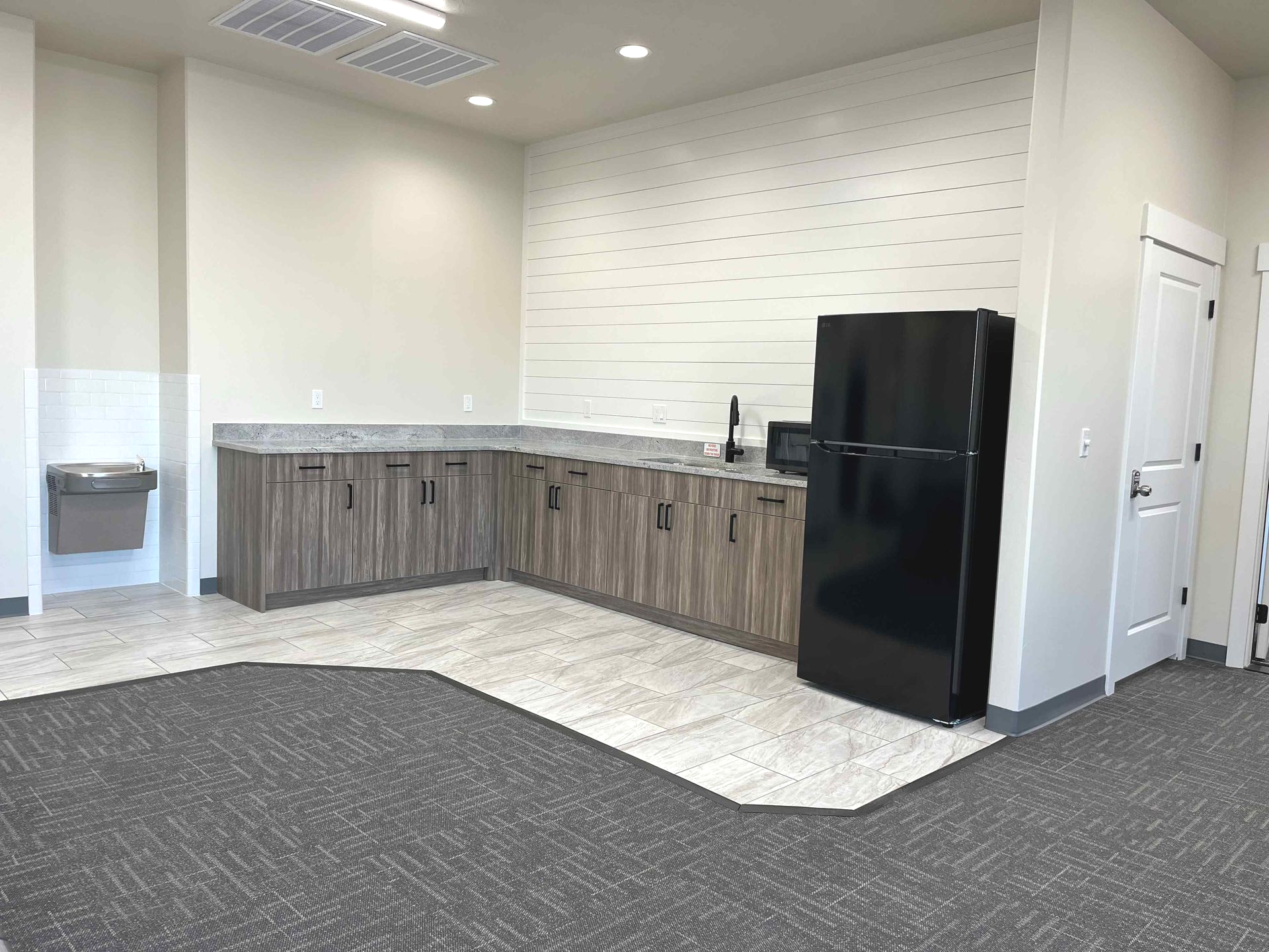 Breakroom with cabinets, granite countertop, black refrigerator, sink, and microwave. Gray carpet and light walls.