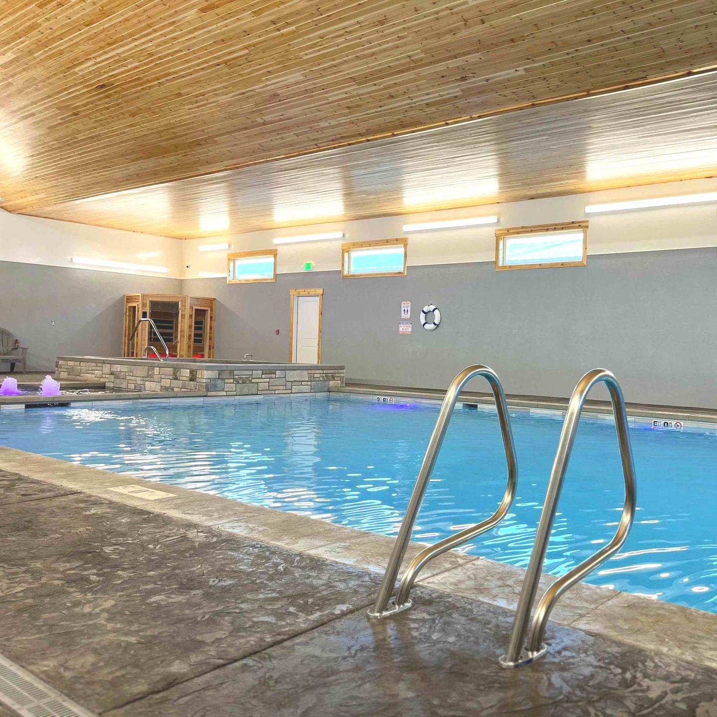 Indoor pool with blue water, stainless steel ladder, light wood ceiling, and sauna in background.