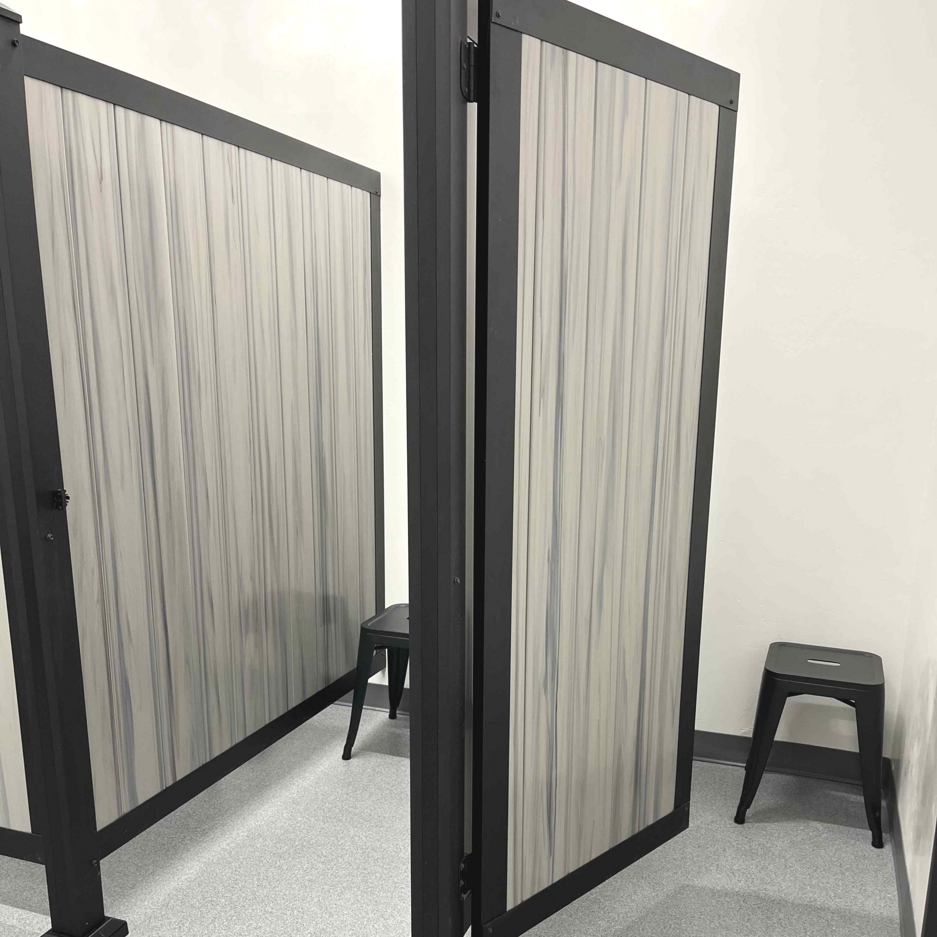 Two gray and black dressing room stalls, each with a stool, set against white walls and a light-colored floor.