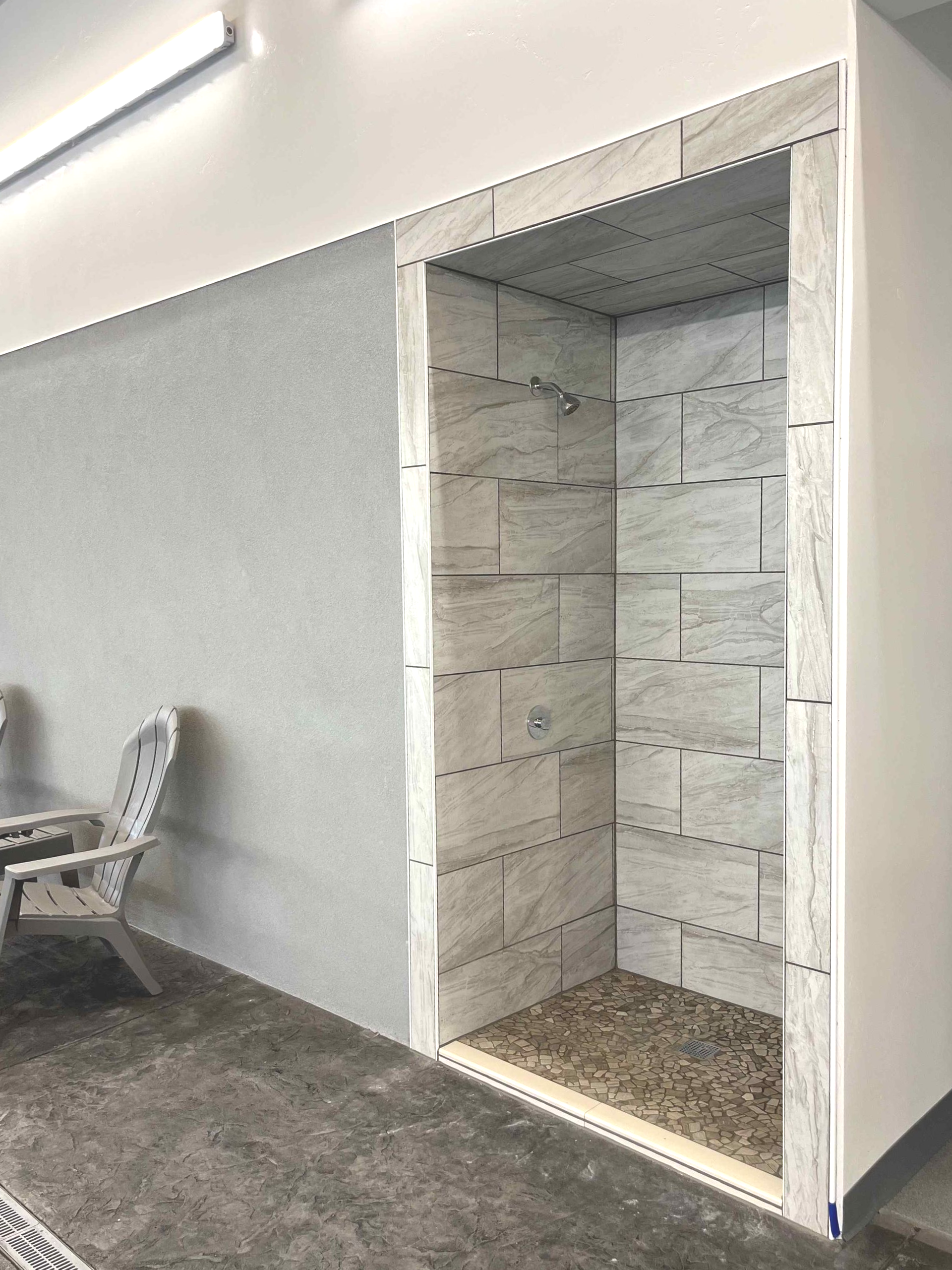 A shower stall with tiled walls and a pebble floor; adjacent to a stucco wall and a chair.