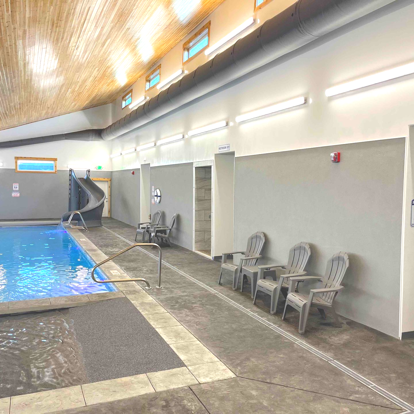 Indoor pool with slide, gray walls, chairs, and a light wooden ceiling.