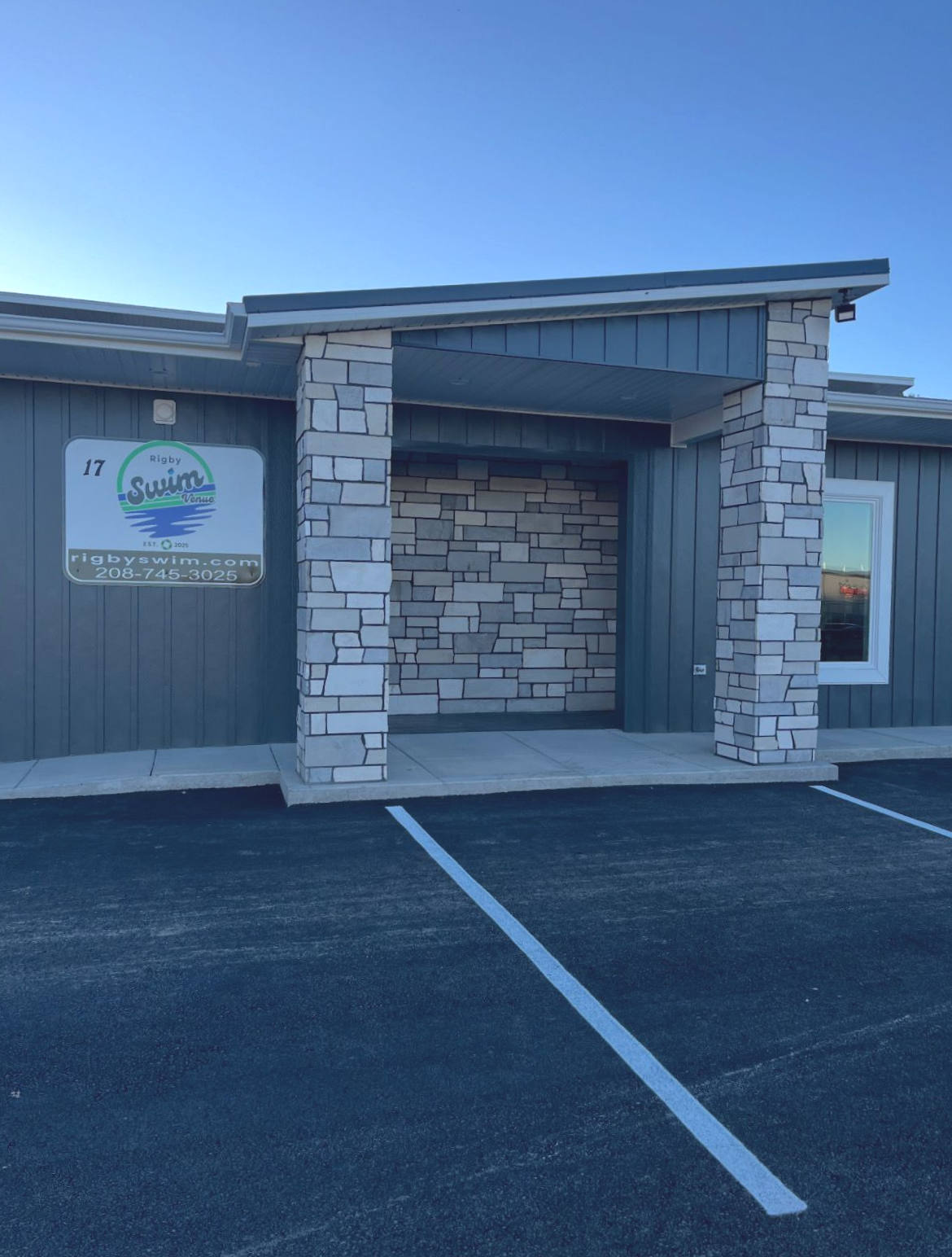 Entrance to a business: Grey building with stone pillars and a sign. Blacktop parking lot.