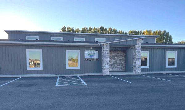 Gray building with stone entrance, windows, and a parking lot on a sunny day.