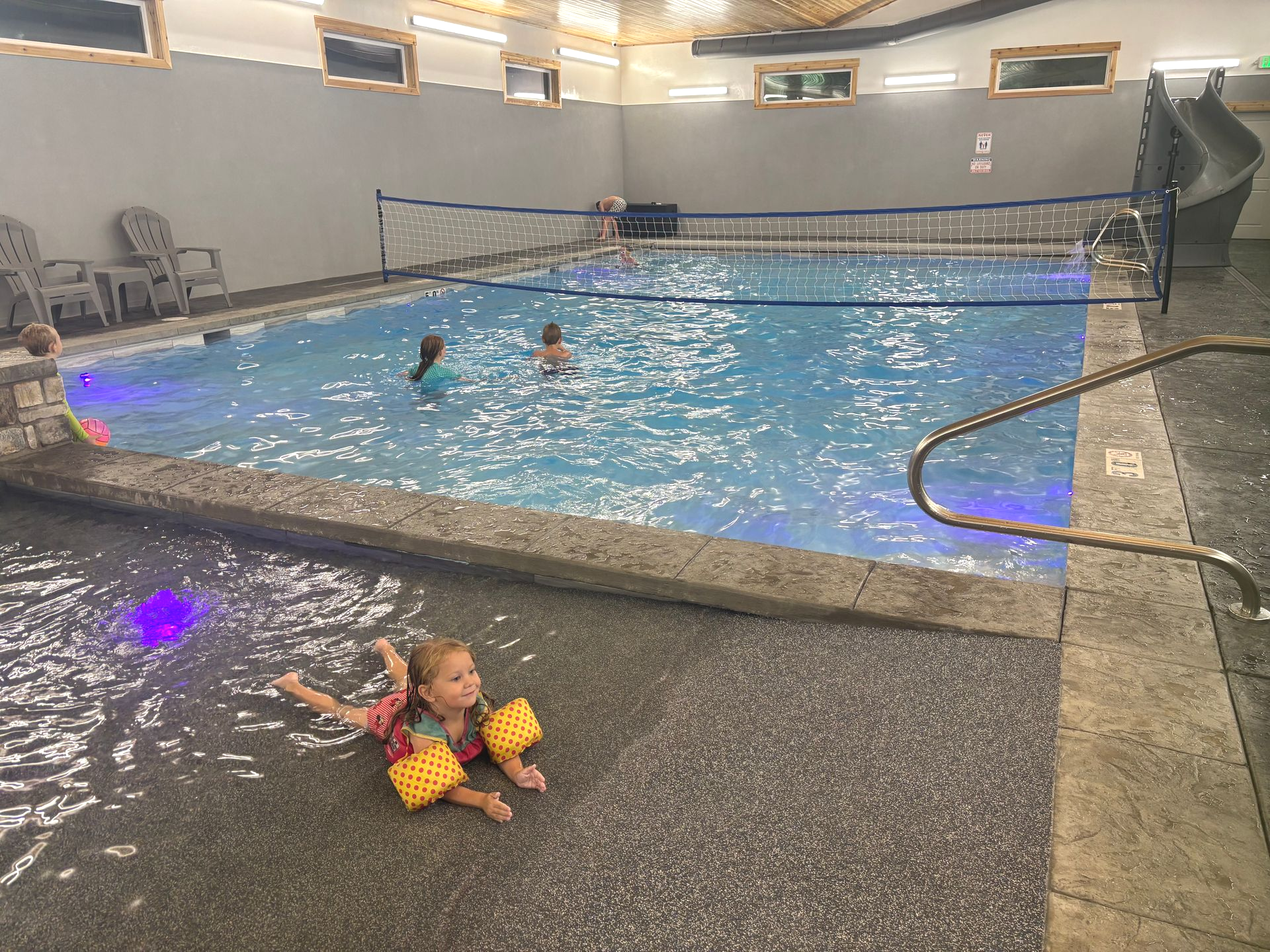 Girl with floaties in pool, others swimming, indoor setting with volleyball net, water slide.