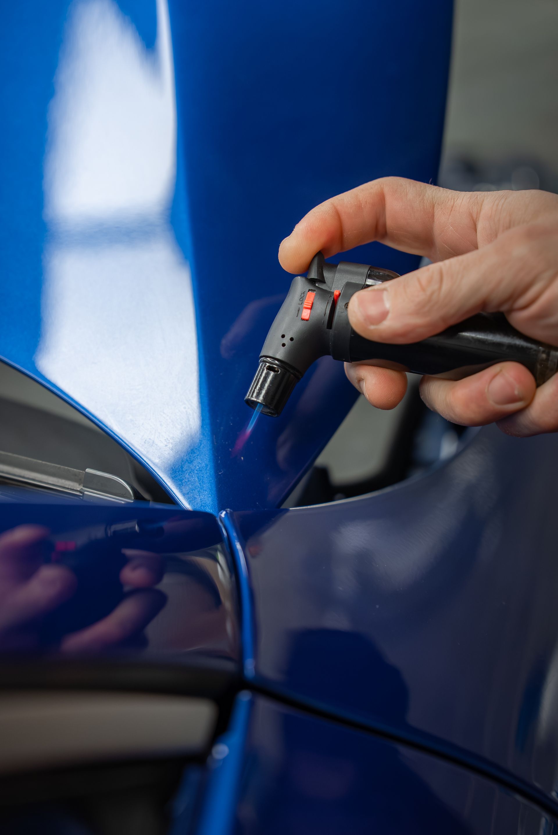 Hand holding a torch over a blue car surface, likely for repair or modification.