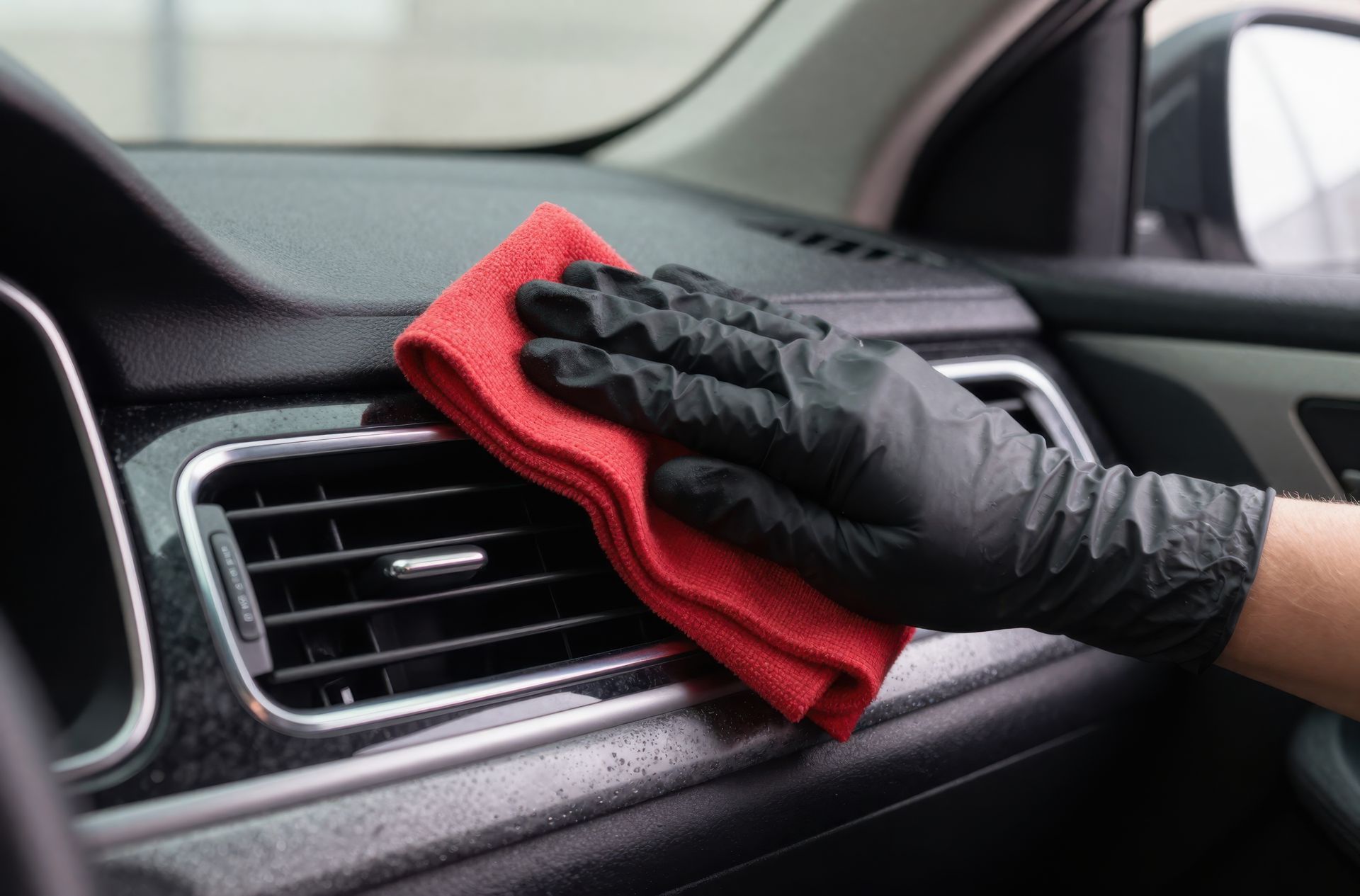 Gloved hand cleaning a car dashboard with a red microfiber cloth.