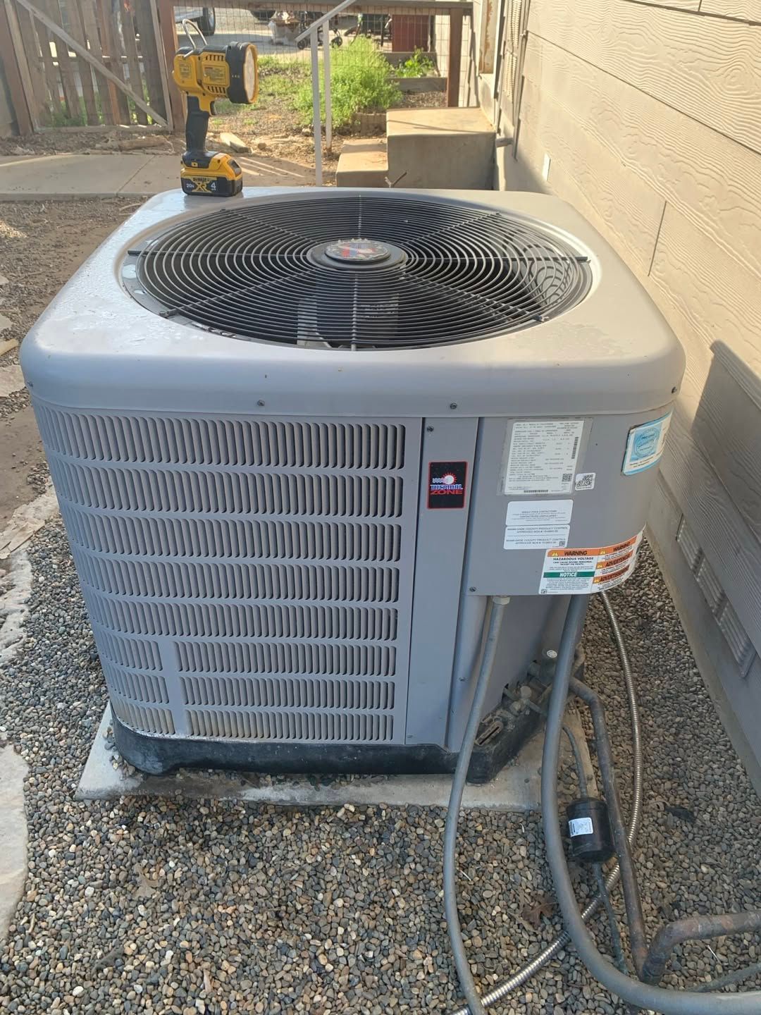 Outdoor air conditioning unit on gravel beside a building, with hoses attached and a large fan on top