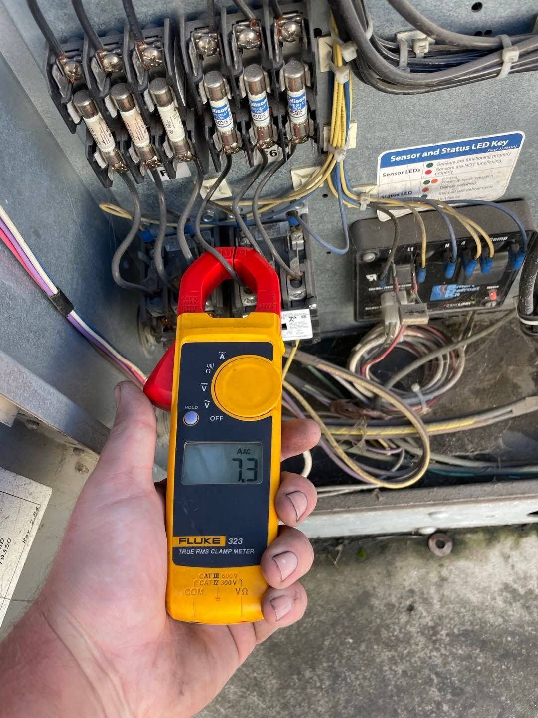 Hand holding a yellow clamp meter inside an open electrical panel with wiring and relays