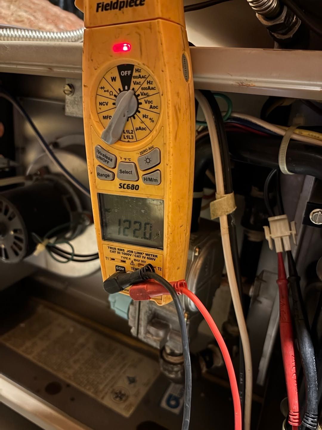 Clamp meter with red probe leads reading 120.0 volts in a cluttered electrical panel area