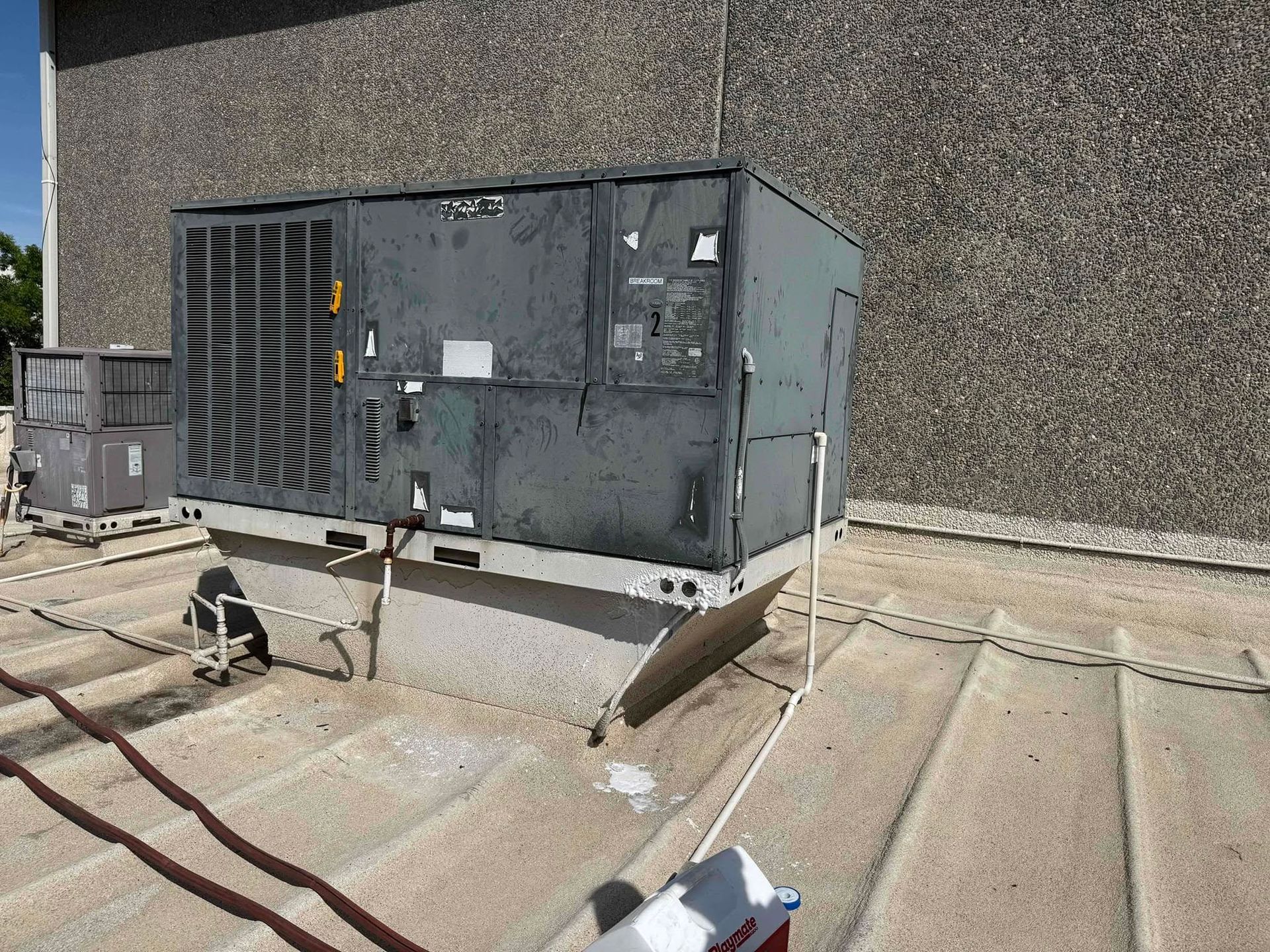 Rooftop HVAC unit beside a wall, with metal vents, pipes, and loose rooftop cables.