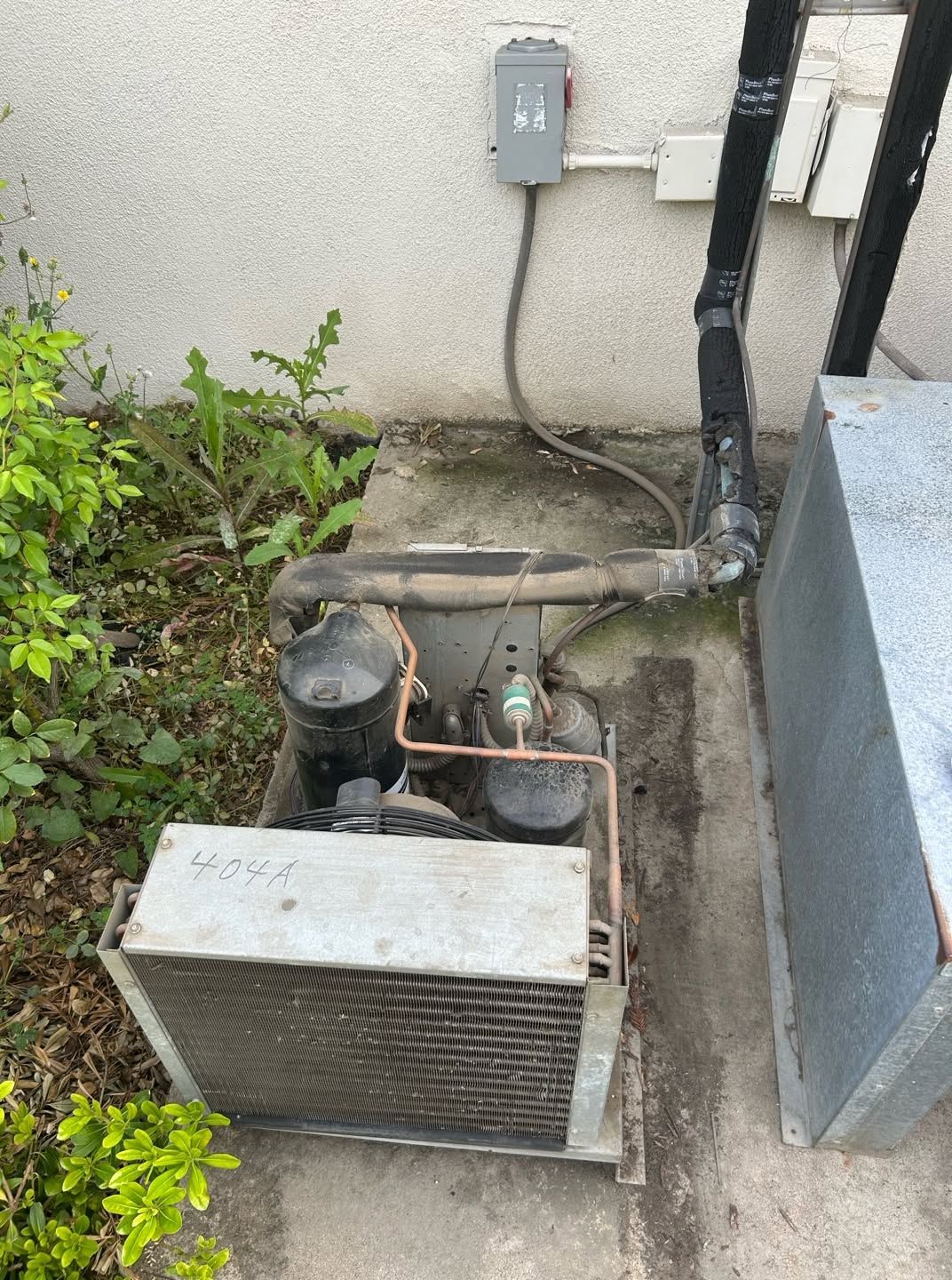 Outdoor HVAC unit beside a wall, with pipes, wiring, and nearby overgrown plants