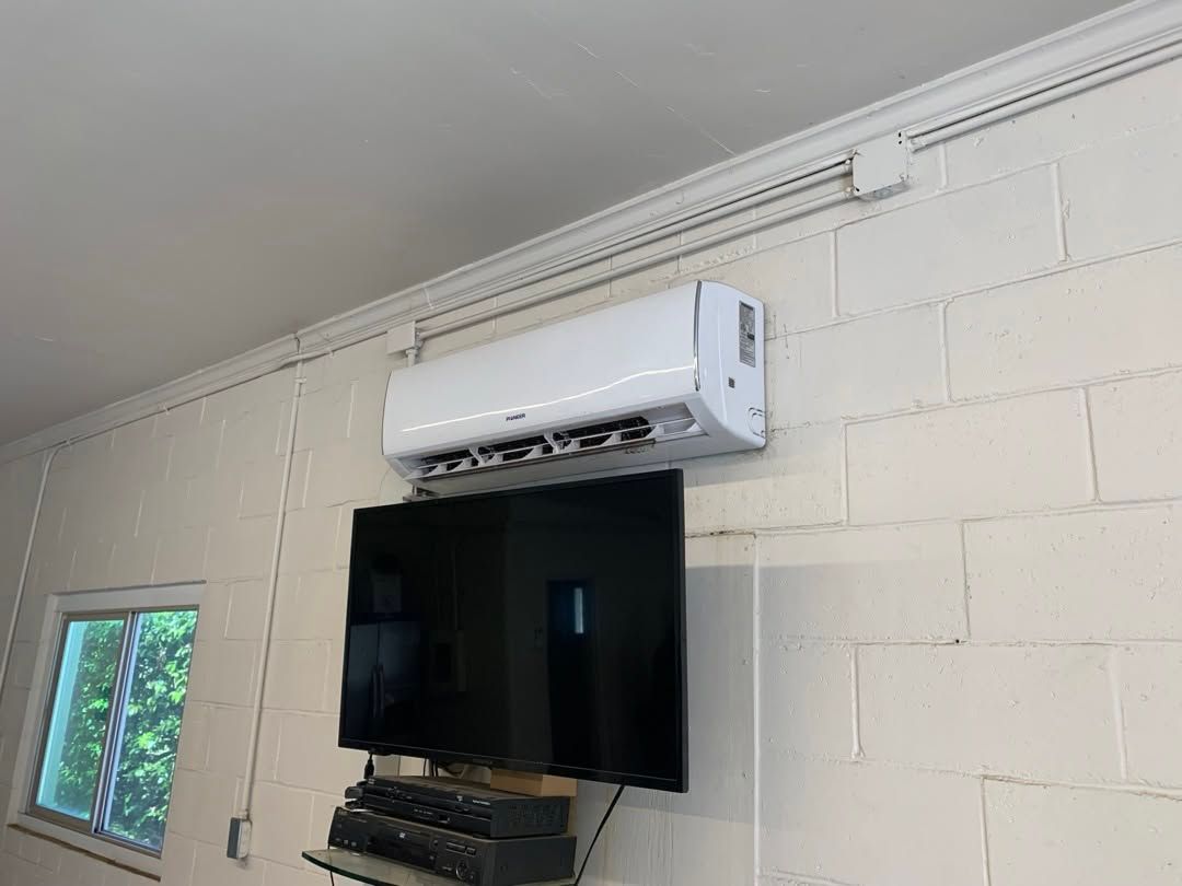 Wall-mounted TV beneath a white mini-split air conditioner in a light-colored room with a window.