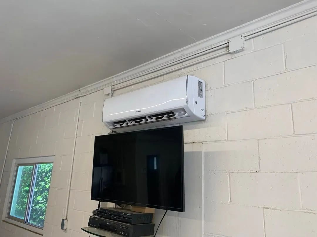 Wall-mounted TV beneath a white air conditioner in a room with cinderblock walls and a window.