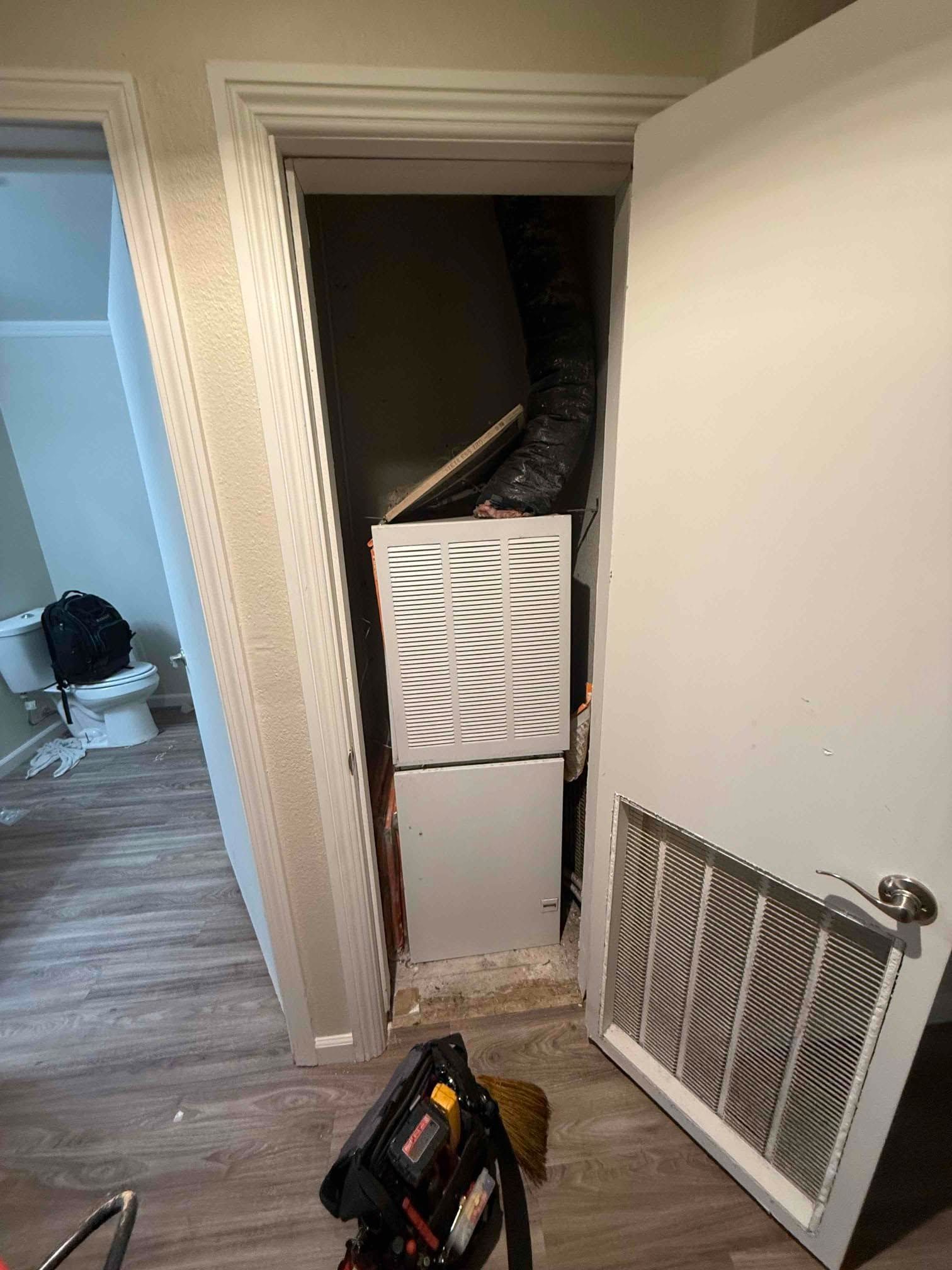 Open utility closet with a white panel leaning below; loose vent cover on floor in hallway.