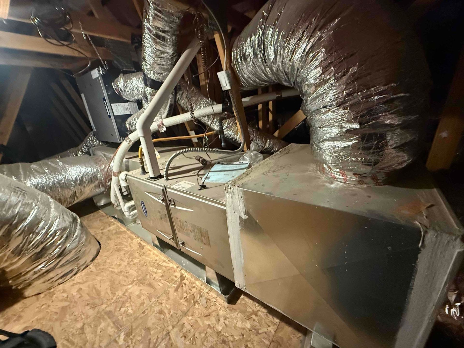 Metal HVAC/furnace unit in an attic surrounded by insulated ductwork and exposed wooden rafters.