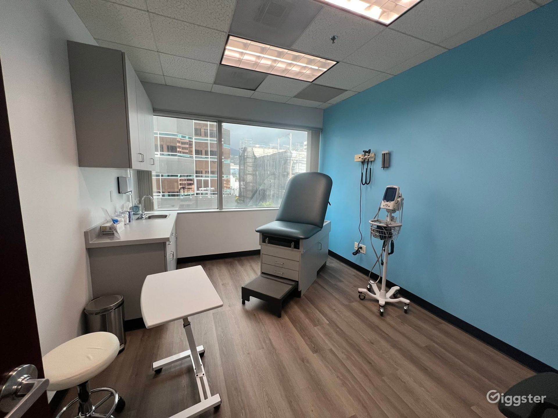 Medical exam room with blue wall, exam chair, stool, and window view of city buildings