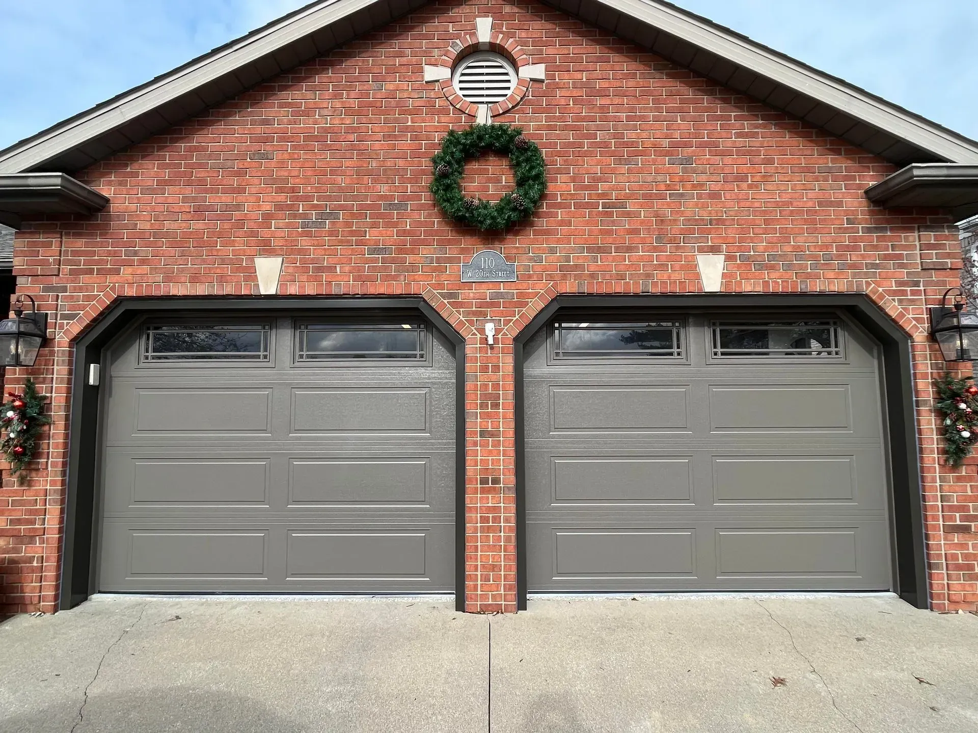 Brick house front with two gray garage doors and a green wreath decoration on the gable