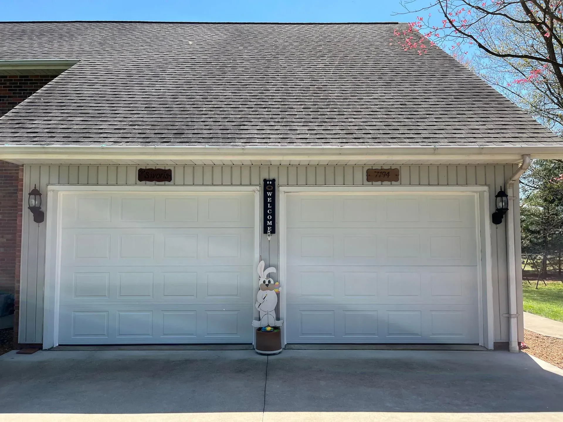 Two white garage doors on a house with a gray shingled roof and brick columns