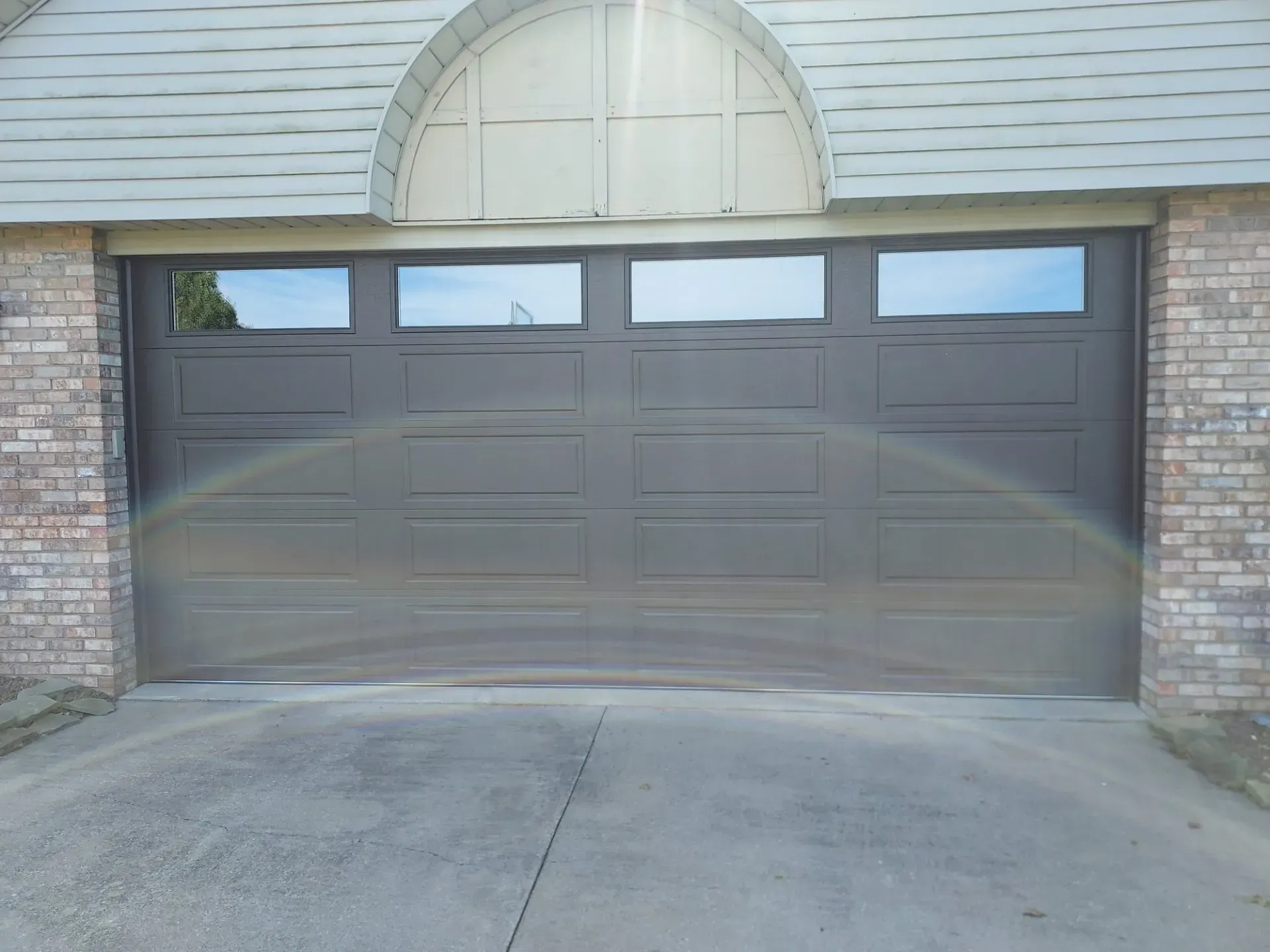 Brown garage door with small windows, framed by brick walls under a light siding arch.