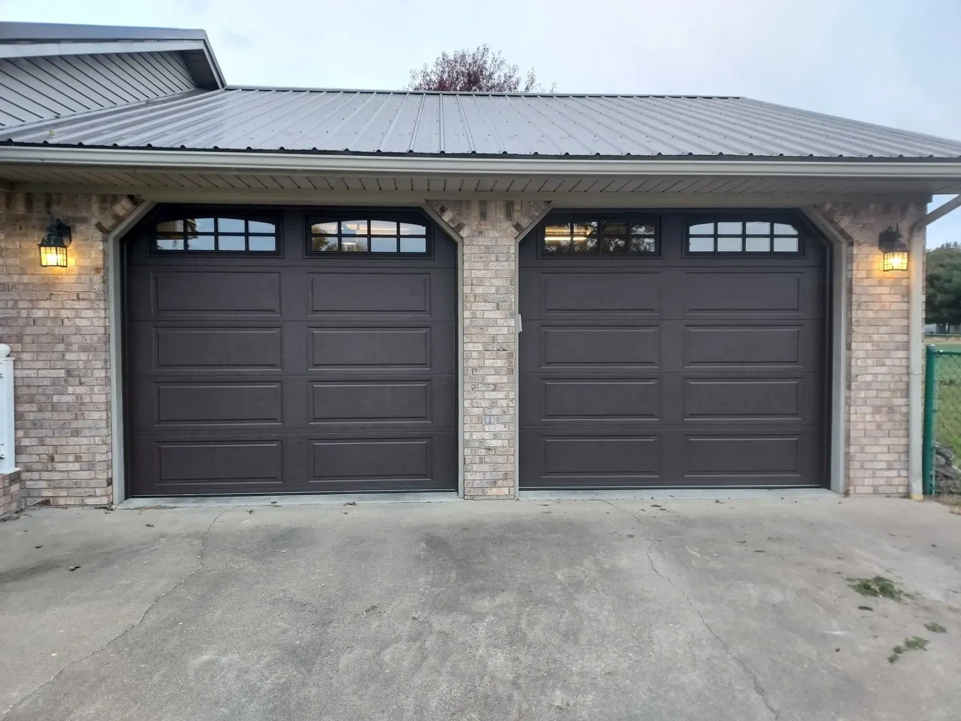 Two dark brown garage doors set in a stone house exterior with a metal roof and driveway