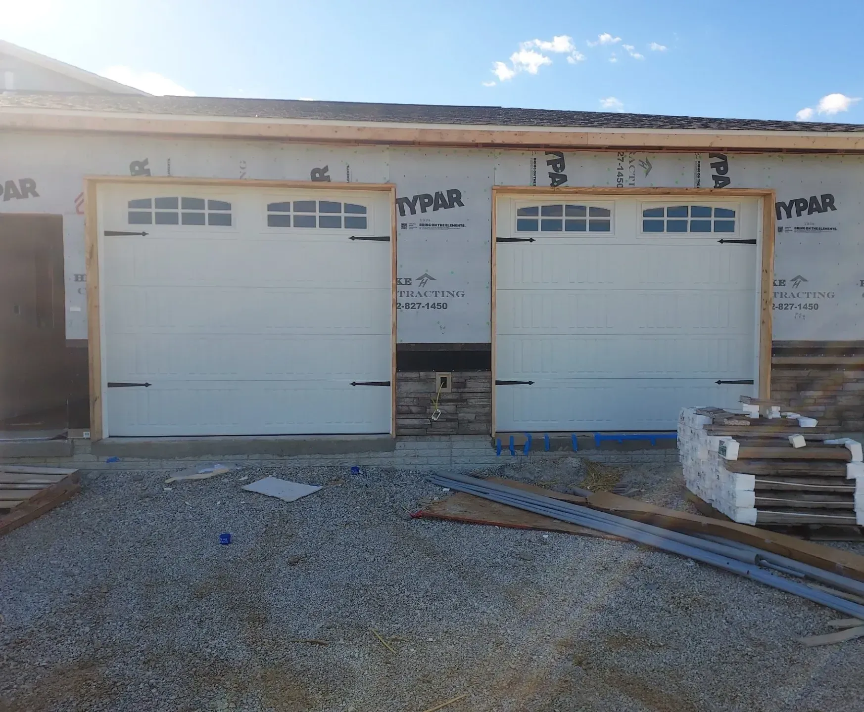 Two white garage doors on a house under construction, with gravel, lumber, and building materials outside.