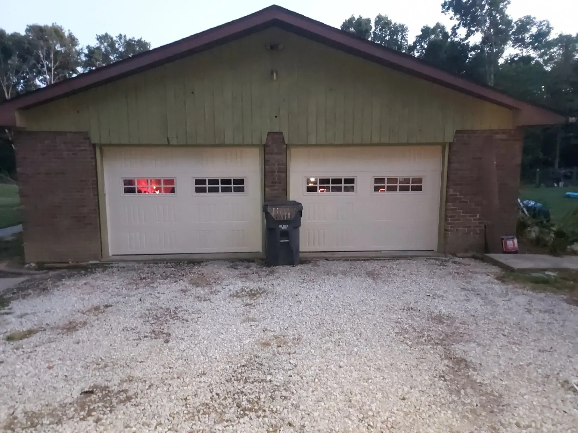 Two-car garage with beige doors, brick columns, and a trash bin on a gravel driveway.