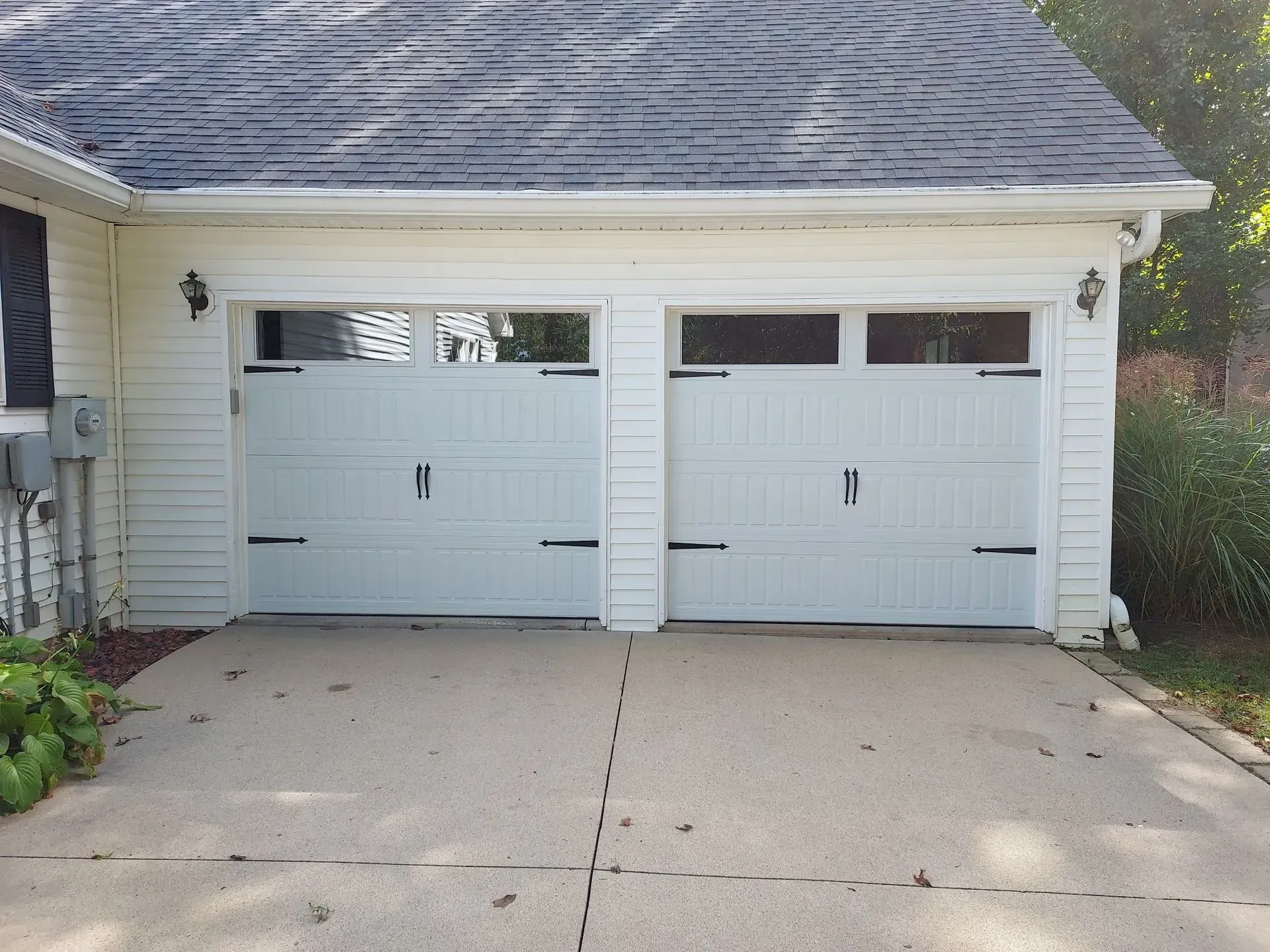 Two white garage doors with small upper windows on a concrete driveway, attached to a house.