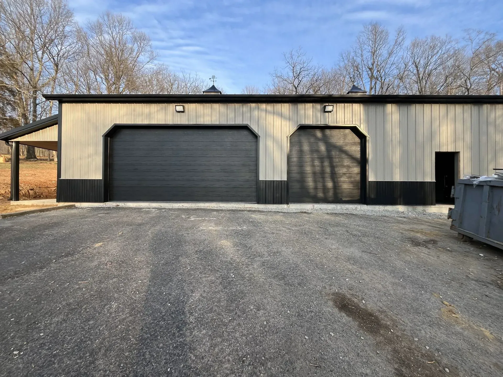 Tan metal building with black garage doors and a gravel driveway under a blue sky