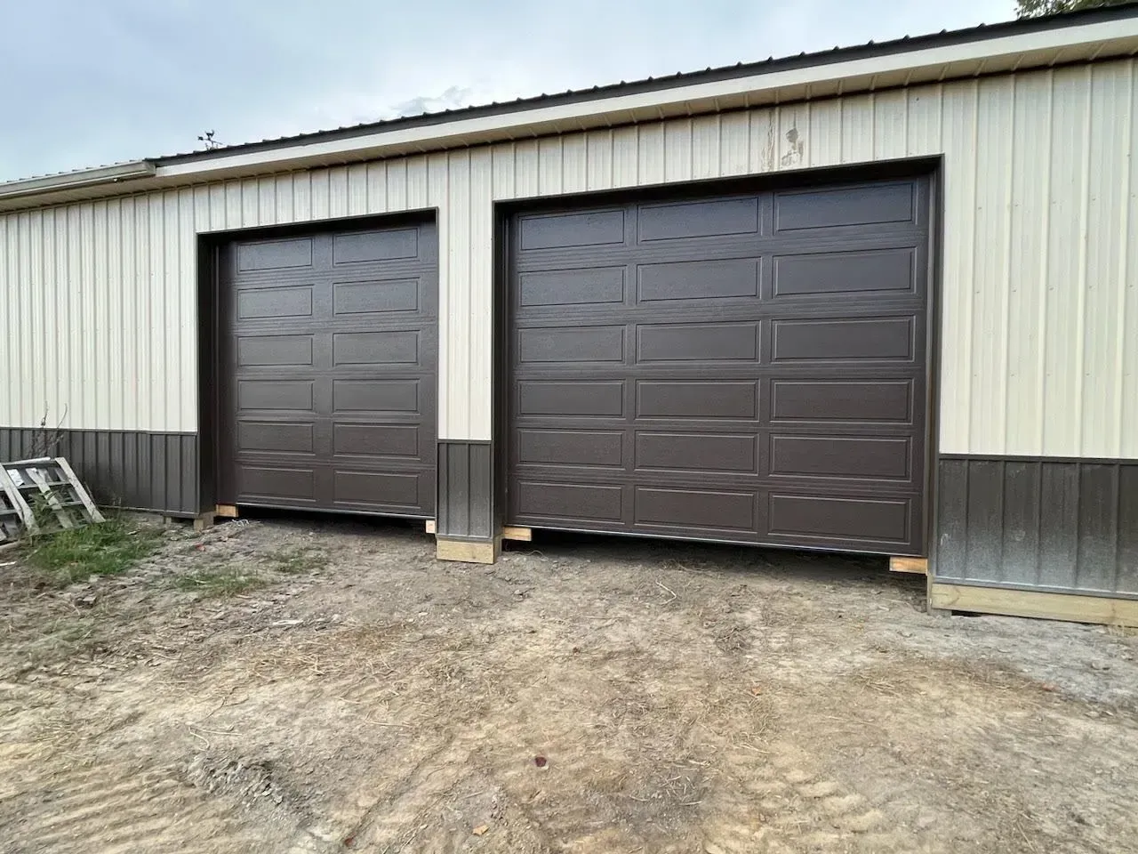 Two dark brown garage doors on a beige metal building over a gravel lot