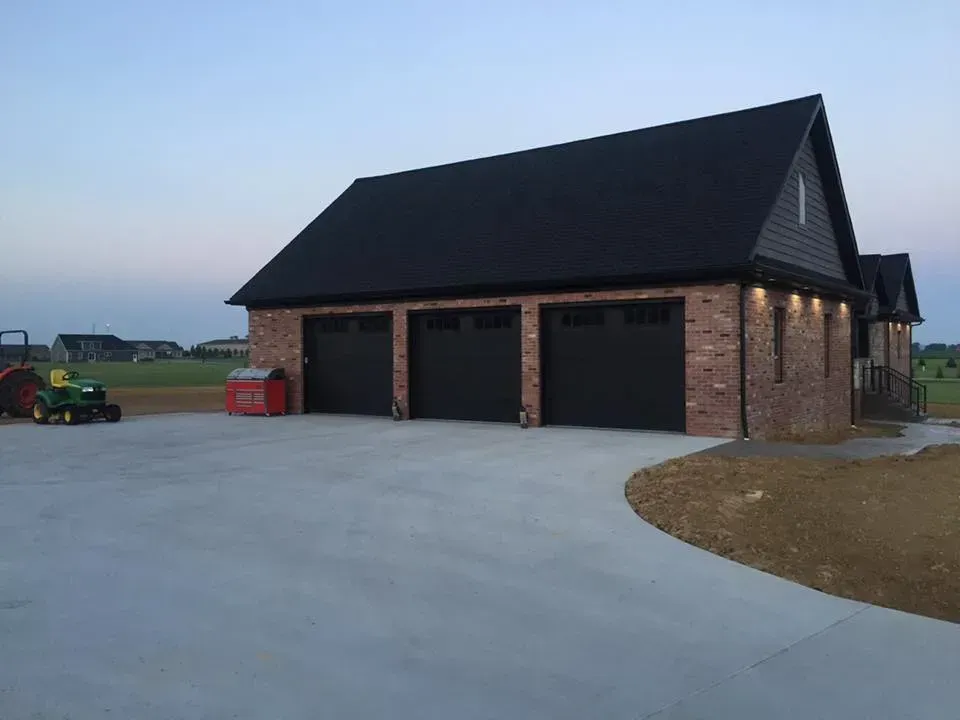 Three-car brick garage with dark roof beside a large concrete driveway at dusk