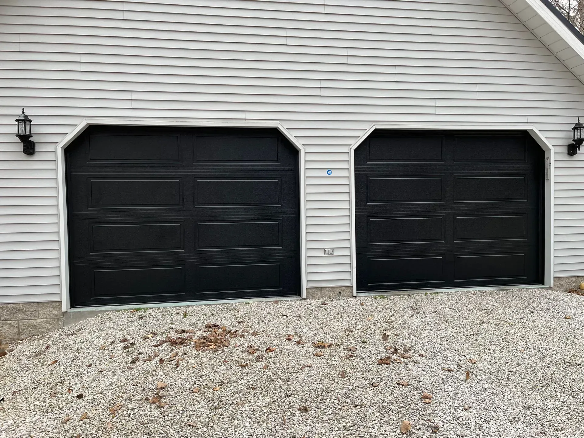 Two black garage doors on a white house with gravel driveway in front