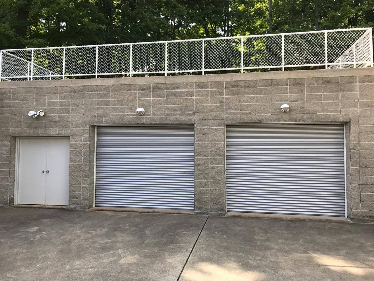 Concrete garage wall with two gray roll-up doors and a small side door, topped by a chain-link fence.
