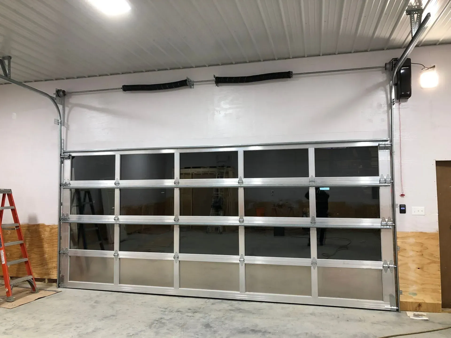 Large gray industrial garage door in a workshop, with a red ladder and wooden wall panels on the left.