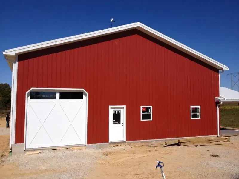 Red barn-style building with white garage door and small entry door on a dirt lot under blue sky