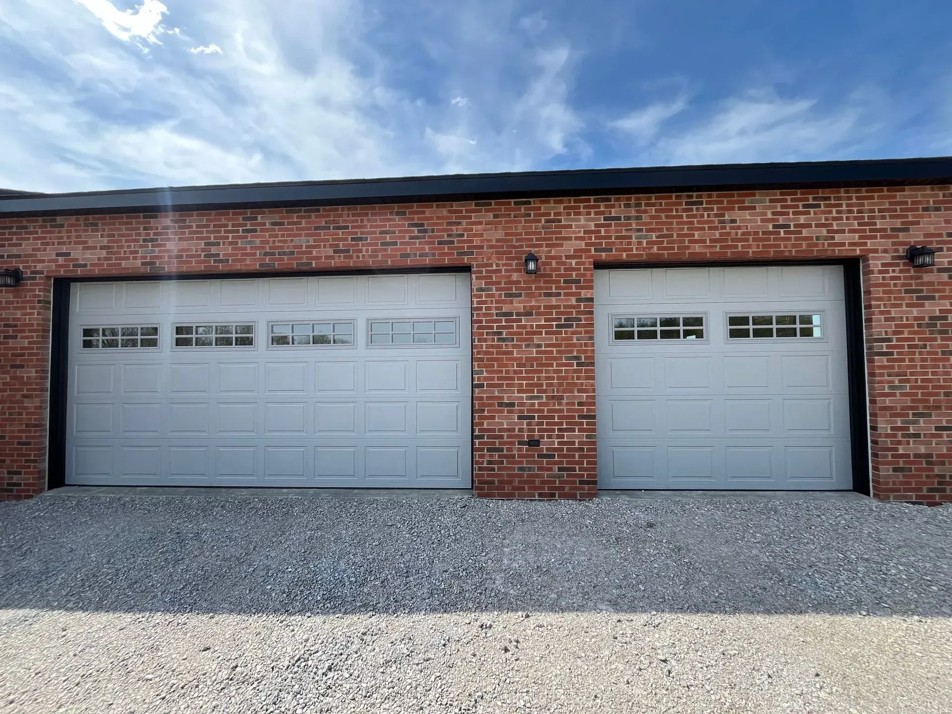 Brick building with two gray garage doors and a gravel driveway under a cloudy sky