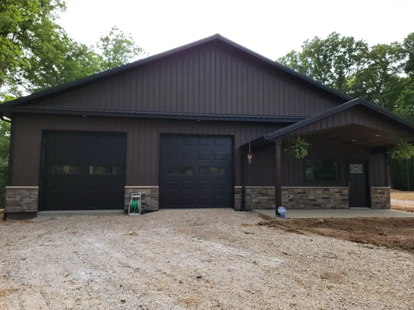 Dark brown house with a large double garage, stone accents, and a gravel driveway amid trees.