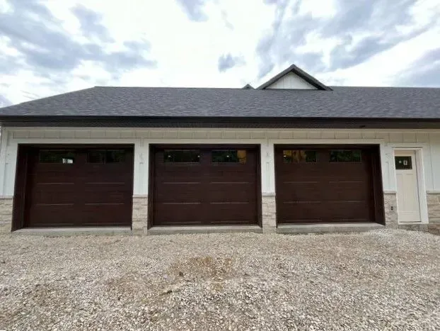 Three brown garage doors on a white house with gravel driveway and cloudy sky