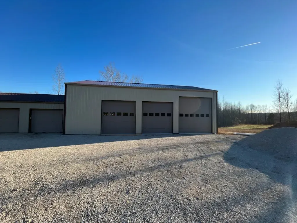 Gravel driveway leading to a beige metal garage with three doors under a clear blue sky.